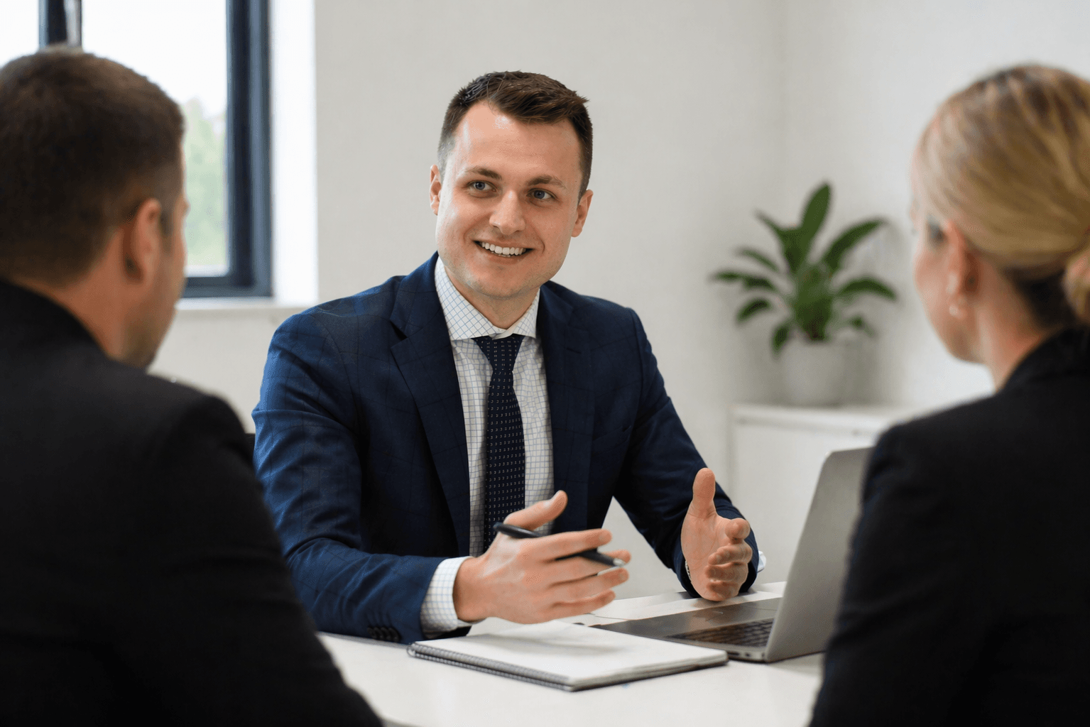 Smiling businessman in a suit gestures while speaking to colleagues during an office meeting.